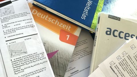 Stapel von Lehrbüchern und Materials liegt verstreut, während jemand für das Homeschooling lernt. In der Umgebung sieht man verschiedene Lernressourcen.