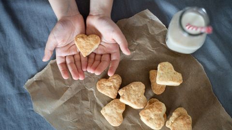 In beiden Händen wird ein herzförmiges Plätzchen gehalten, umgeben von weiteren Plätzchen auf einer papierten Unterlage, neben einem Glas Milch.