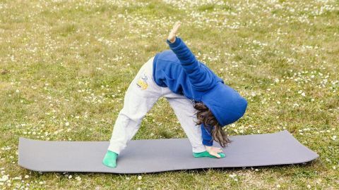 Ein Kind übt eine Yoga-Position auf einer Matte in einer blühenden Wiese, umfit mit einem blauen Hoodie und weißen Hosen.