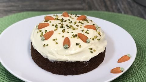 Ein saftiger Karottenkuchen steht auf einem weißen Teller, dekoriert mit Karotten und Pistazien, auf einem grünen Platz.