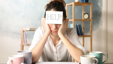 Eine Person sitzt an einem Tisch, den Kopf in den Händen und hält ein Schild mit "HELP" hoch, um Unterstützung in stressigen Zeiten zu signalisieren.