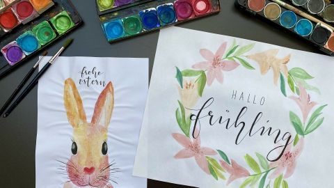 Bunte Wasserfarben liegen neben zwei Bildern: eines eines Hasen, das andere mit dem Schriftzug "Hallo Frühling".