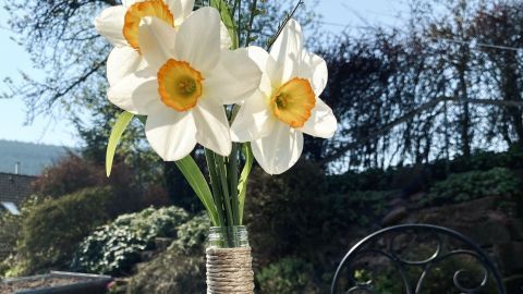 Eine mit Jute-Schnur umwickelte Flasche steht als Vase mit weißen Narzissen auf einem Tisch im Garten.