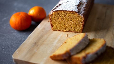 Ein Mandarinenkuchen liegt auf einem Holzbrett, während zwei frische Mandarinen daneben sind und einige Scheiben abgeschnitten wurden.