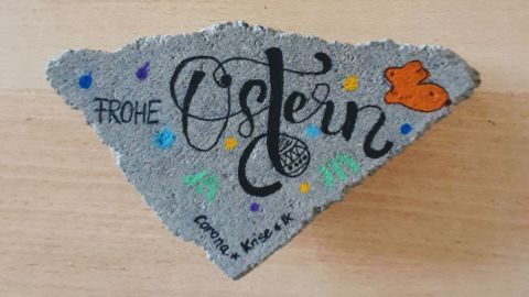 Ein grauer Stein zeigt die farbenfrohe Aufschrift "Frohe Ostern" mit bunten Verzierungen und einem Schmetterling.