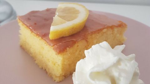 Ein saftiges Stück Zitronenkuchen liegt auf einem rosa Teller, garniert mit einem Zitronenschnitz und Sahne.
