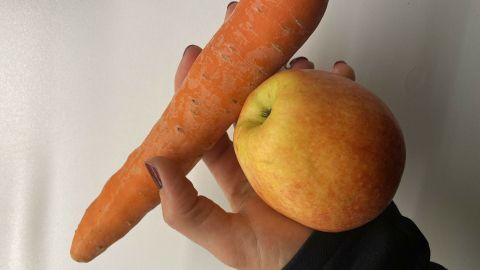 Eine Hand hält eine Karotte und einen Apfel als gesunde Snack-Alternativen in einer hellen Umgebung.