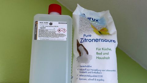 Eine Flasche mit Desinfektionsmittel steht neben einem Beutel Zitronensäure, beide auf einem hellgrünen Hintergrund.