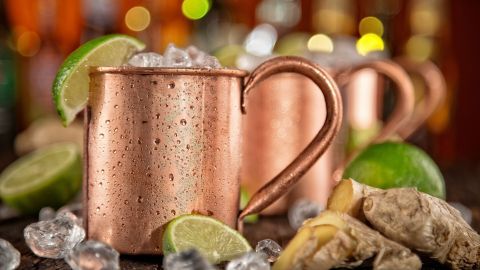 Kupferne Becher mit Eis, Limettenscheiben und Ingwer stehen auf einem Holztisch in einer barähnlichen Umgebung.