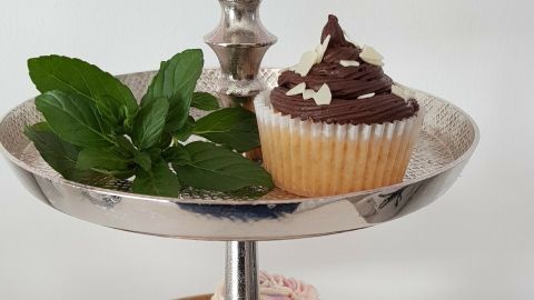 Auf einem eleganten Etagere stehen mehrere Vanille-Muffins, dekoriert mit Schokolade und Sahne, umgeben von frischen Blättern.