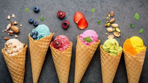 Sechs Eiskugeln in Waffeln stehen auf einem dunklen Untergrund, umgeben von Beeren und Nüssen, zeigen die Vielfalt von Speiseeis.