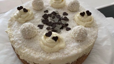 Ein Raffaello-Kuchen, dekoriert mit Sahne und Schokoladenherzen, steht auf einem Teller in einer hellen Umgebung.