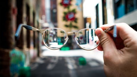 Eine Hand hält eine Brille hoch, während die Straße im Hintergrund klar und belebt ist; Tipps zur Vermeidung von beschlagenen Brillengläsern sind relevant.