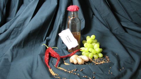 Eine Glasflasche mit einer Weißweinreduktion steht vor grünem Trauben- und Chiligemüse, auf einem dunkelblauen Stoff.