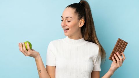 Eine Frau hält einen Apfel in der einen Hand und eine Schokolade in der anderen, während sie lächelt und entspannt vor einer blauen Wand steht.