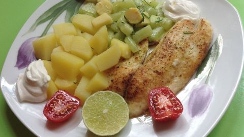 Ein gebratenes Tilapia-Fischfilet liegt auf einem Teller, umgeben von Salzkartoffeln, Gurken und frischen Tomaten.