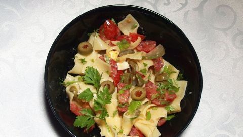 Der Sud von eingelegten Peperonis eignet sich sehr gut, um Salatsoßen zu verfeinern. Natürlich muss sparsam dosiert werden. Ein schwarzes Schüsselchen mit Salat ist gefüllt mit Nudeln, Tomaten, Oliven und frischen Kräutern, dekoriert mit Peperonisud.