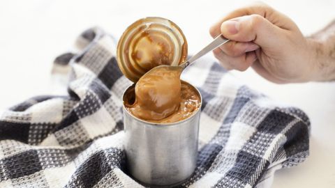 Eine Hand schöpft einen glänzenden Löffel mit selbstgemachtem Dulce de leche aus einer geöffneten Dose, umgeben von einem karierten Tuch.