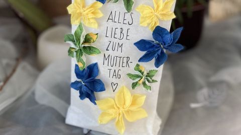 Eine dekorative Lichttüte ziert mit bunten Papierblüten den Schriftzug „ALLES LIEBE ZUM MUTTERTAG“ im Raum.