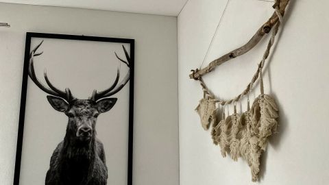 Eine dekorative Makramee-Feder hängt an einer Wand, daneben ein großes Schwarz-Weiß-Bild eines majestätischen Geweihs.