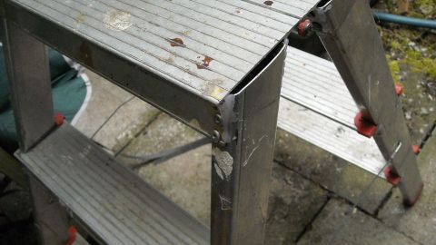 Ein Haushaltsleiter steht schräg auf den Bänken und wirkt instabil, umgeben von einem schlichten Außenbereich.