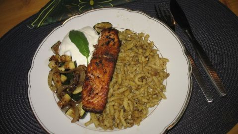 Ein gebratenes Lachsfilet liegt auf einem Teller mit Zucchinigemüse und Eierspätzle, angerichtet auf einem gedrungenen Tisch.