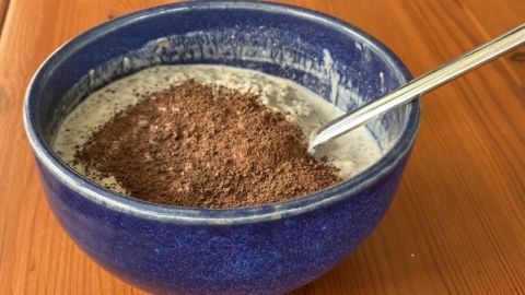 Eine blaue Schüssel mit Müsli steht auf einem Holztisch, während ein Löffel im Müsli steckt, das mit Schokoladenpulver bestreut ist.