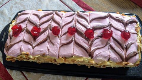 Ein appetitlich dekorierter Esterhazyschnitt mit Buttercreme und roten Kirschen liegt auf einem schwarzen Blech.