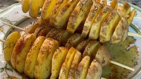 Zwei Spieße mit spiralförmig geschnittenen, gewürzten Kartoffeln liegen auf einem Teller, bereit zum Grillen.