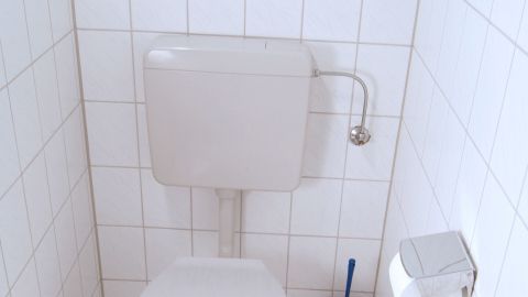 Eine Toilette mit Spülkasten, die ständig läuft, steht in einem gefliesten Raum. Neben ihr befindet sich eine Toilettenbürste und Toilettenpapier.