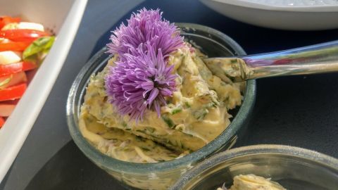 Ein Glas mit Schnittlauchblütenbutter, in dem eine lila Blüte dekorativ platziert ist, steht auf einem Tisch neben frischem Gemüse.