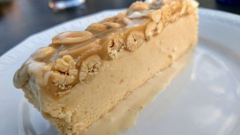 Ein Stück Snickers-Eis liegt auf einem weißen Teller. Es ist schaumig und garniert mit Erdnüssen, umgeben von einem klaren Glas Getränk.