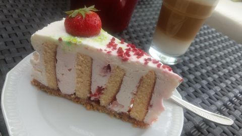 Eine elegante Erdbeer-Holunderblüten-Wickeltorte liegt auf einem weißen Teller, neben einem Glas Latte, auf einem strukturierten Tisch.
