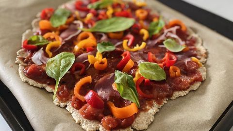 Eine Haferflocken-Pizza liegt auf einem Backblech mit buntem Gemüse und frischen Kräutern, bereit zum Backen.