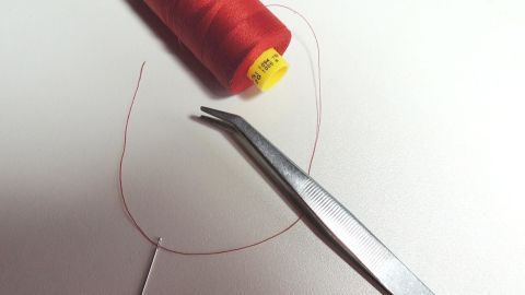 Eine rote Nähgarnrolle liegt neben einer Zange, die dabei ist, den Faden einfach und schnell einzufädeln.
