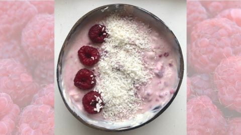Ein cremiger Kokos-Himbeer-Joghurt in einer Schüssel, garniert mit Himbeeren und Kokosraspeln, vor rosa Hintergrund.