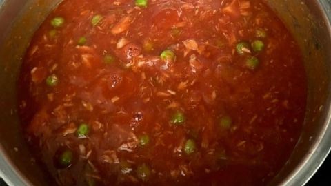 Eine rote Nudelsoße mit Thunfisch und Erbsen kocht in einem Topf auf dem Herd, umgeben von einer modernen Küchenumgebung.