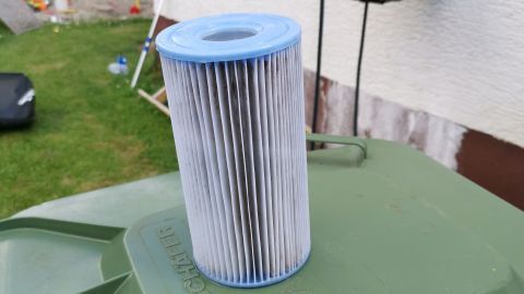 Ein runder Poolfilter steht auf einem grünen Abfallcontainer, umgeben von Rasen und Bäumen, bereit zur Reinigung.