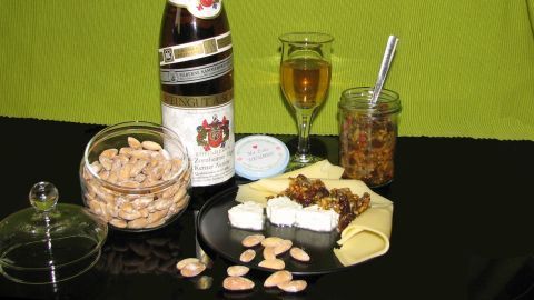 Eine Flasche Agrodolce Sauce steht auf einem schwarzen Tisch, umgeben von Mandeln, einem Glas, Käse und einem Marmeladenglas.