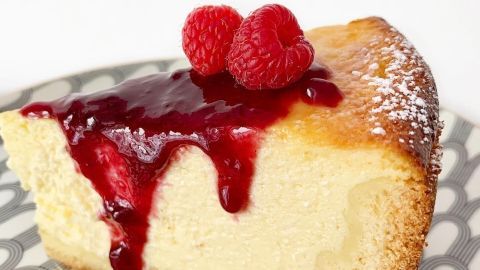 Ein saftiger Käsekuchen mit roter Fruchtsauce und frischen Himbeeren wird auf einem dekorativen Teller präsentiert.