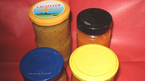 Verschiedene Gläser mit Marmelade ohne Gelierzucker stehen auf einer roten Tischdecke. Die Farben der Marmelade variieren.