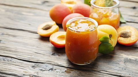 Gläser mit Aprikosenmarmelade stehen neben frischen Aprikosen und Zuckermelonen auf einem rustikalen Holzuntergrund.