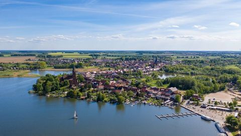 Eine malerische Landschaft mit einem See, auf dem ein Segelboot fährt, umgeben von üppigem Grün und einer kleinen Stadt.