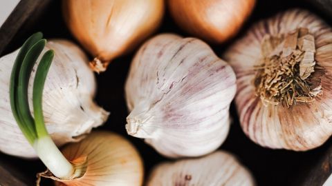 Eine Holzkiste mit verschiedenen Zwiebeln und Knoblauch liegt auf einem hellen Tisch, während die Zutaten frisch und einladend wirken.
