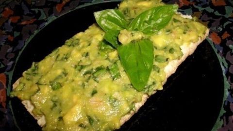 Ein knuspriges Brotstück mit Avocado und Basilikum liegt auf einem schwarzen Teller, umgeben von einer bunten Dekoration.