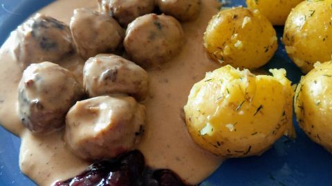Saftige schwedische Köttbullar liegen auf einem blauen Teller mit Sahnesauce und Beilagen aus Kartoffeln und Preiselbeeren.
