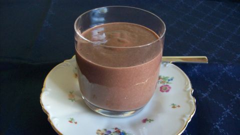 Ein Glas mit brauner, cremiger Hollandse Vla steht auf einem zierlichen Teller in einer eleganten Umgebung.
