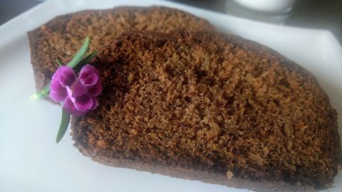 Ein Stück Bananenkuchen liegt auf einem weißen Teller, verziert mit einer kleinen Blume, in einer gemütlichen Umgebung.