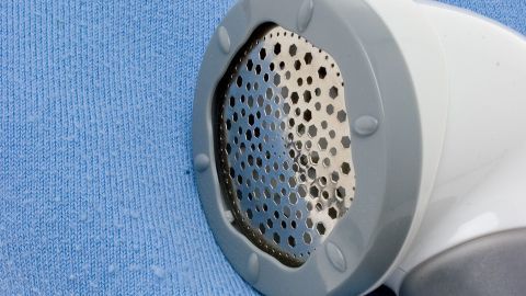 Mit einem einfachen Fusselrasierer, kannst du Knötchen auf der Couch sehr gut entfernen. Ein Fusselrasierer entfernt Knötchen von einem blauen Pullover, der auf einem hellen Untergrund liegt.