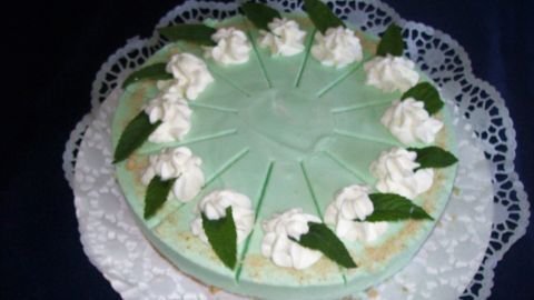 Eine grüne Kühlschranktorte mit Waldmeister-Sahne, dekoriert mit Sahnetuffs und Minzblättern, steht auf einer Spitze.